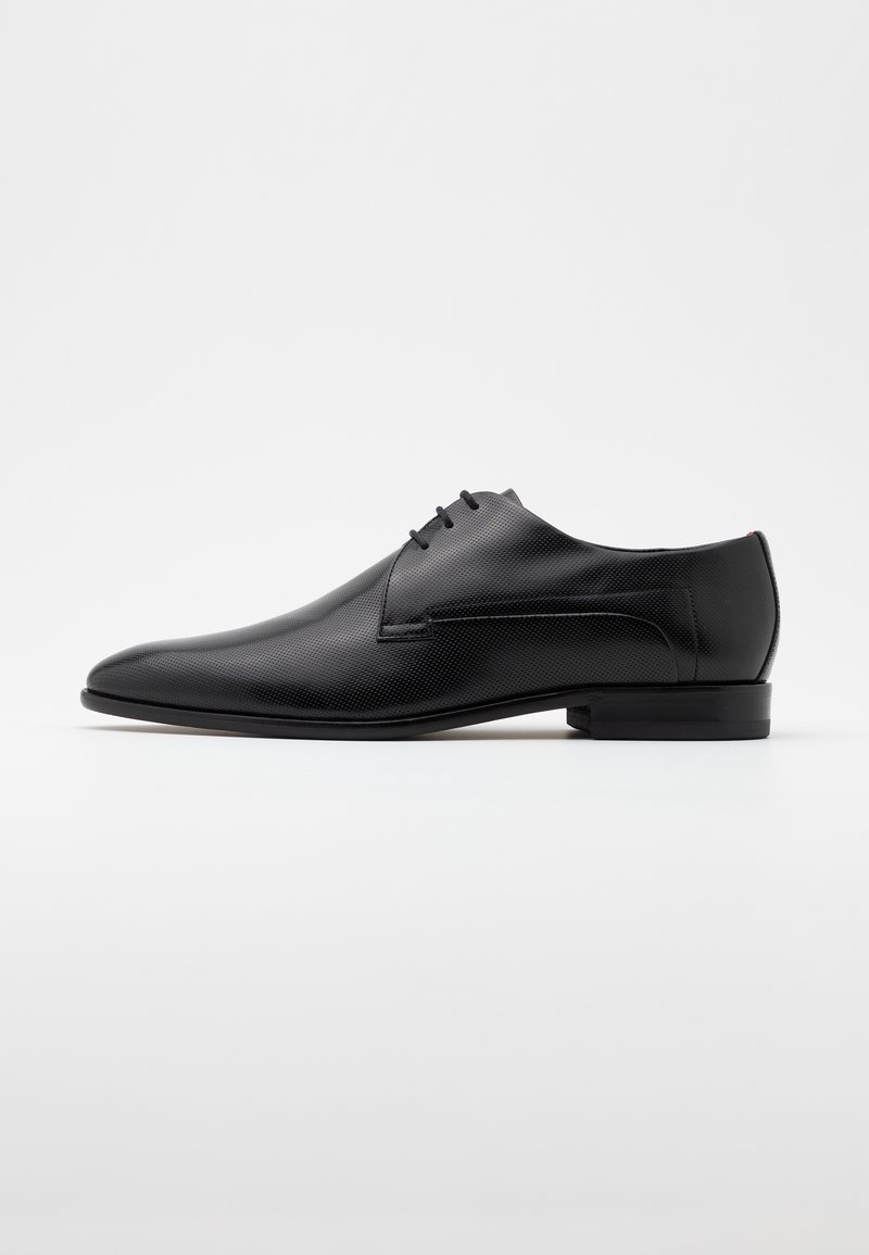 Chaussure de ville pour homme en cuir texturé noir, avec bout pointu et trois œillets pour lacets, sur un fond clair uni.