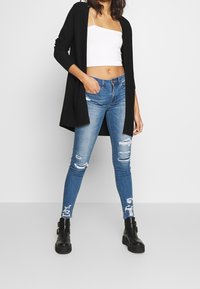 Svart långärmad cardigan över en vit kortärmad topp, i kombination med slitna skinny jeans i blå denim, med revor och svarta ankelboots.