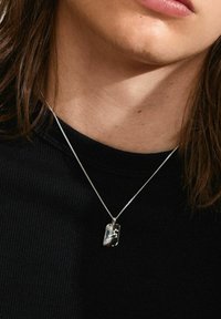 Collarín de plata sobre una cadena delgada, con forma rectangular y acabado pulido, con un sutil grabado, usado sobre una camiseta negra de canalé.