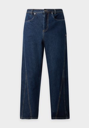 Mand stående iført en kortærmet blå og hvidstribet sweater, mørkeblå jeans og sorte snørestøvler på en ensfarvet baggrund.