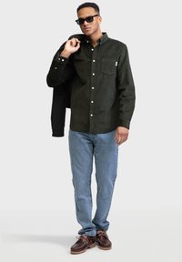 Donkergroen overhemd met knopen en een borstzak, gecombineerd met lichtblauwe jeans en bruine schoenen. Het overhemd heeft een zachte textuur.
