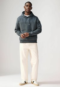 Donkergrijze hoodie met een gestructureerd bovenpaneel, witte trekkoorden, in combinatie met crèmekleurige broek en sneakers met zwarte accenten.
