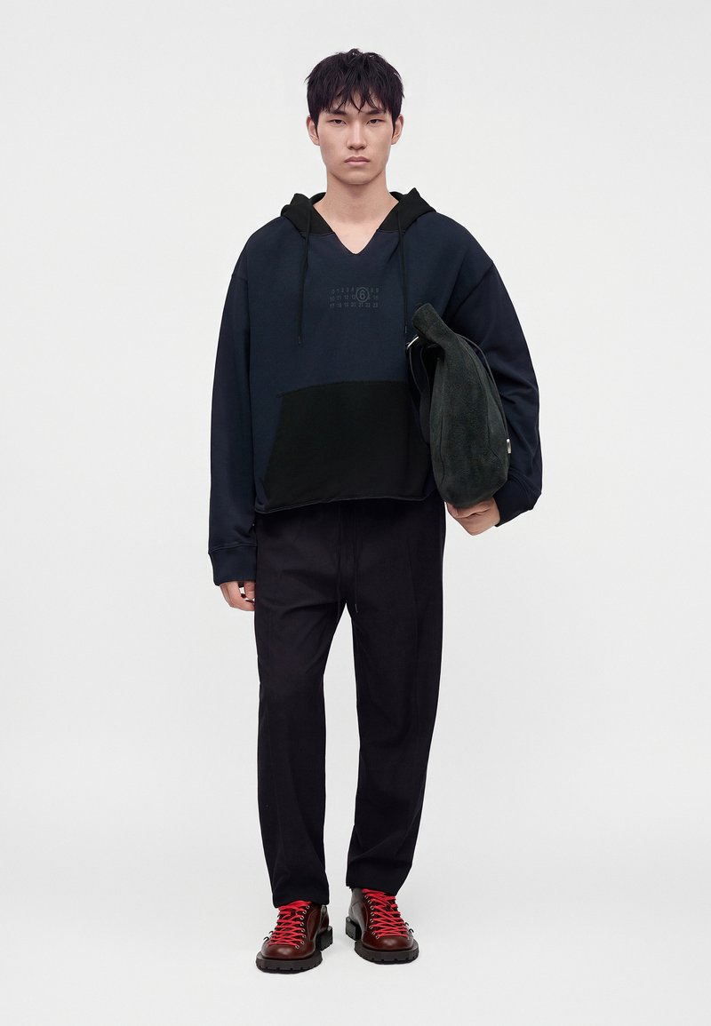 Giovane uomo che indossa una felpa nera con cappuccio, pantaloni neri e stivali marroni con lacci rossi, che tiene una borsa nera con texture su uno sfondo bianco semplice.