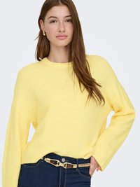 Suéter de punto amarillo con cuello redondo y mangas largas, combinado con jeans oscuros que llevan un cinturón estampado. Textura suave, diseño casual.