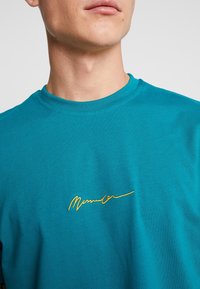 Detailní záběr na osobu, která má na sobě tyrkysové tričko s kulatým výstřihem a žlutým vyšitým kurzívním textem na hrudi.