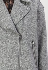 Veste en tricot grise avec une finition texturée, dotée d'un col à revers, d'une fermeture éclair sur le devant et de poches latérales, ainsi que d'accents en métal argenté.