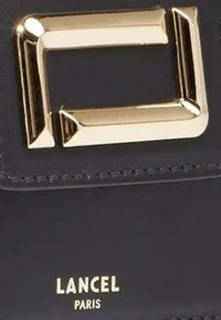 Accessoire en cuir noir avec un accent en métal rectangulaire doré poli et le logo en relief "LANCEL PARIS". Lignes épurées et texture minimale.