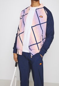 Värikäs bomberjacka med en gradientdesign i rosa och lila, marinblå ärmar, geometriskt mönster och logotypdetaljer. Matchas med marinblå byxor.