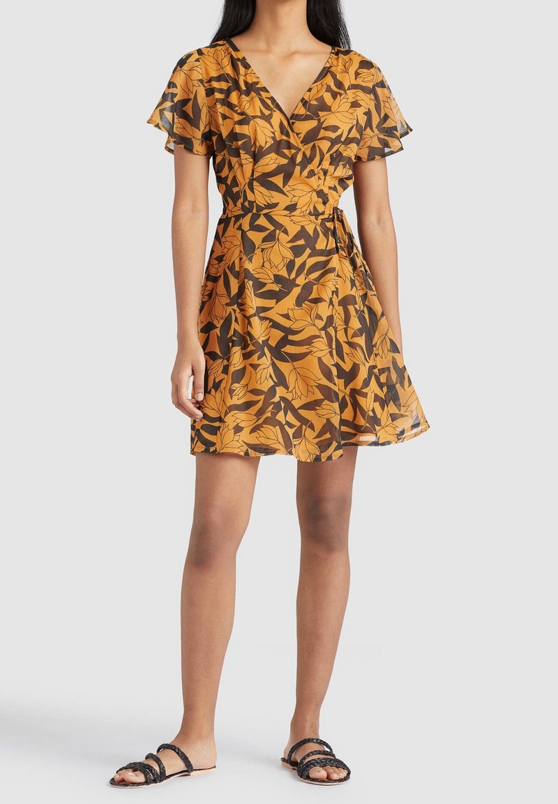 Femme portant une courte robe portefeuille orange avec des motifs de feuilles noires et des manches volantées, associée à des sandales plates noires tressées.