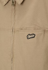 Chaqueta beige con cremallera hecha de una tela resistente. Presenta un cuello puntiagudo, un bolsillo frontal y un logo bordado en negro en el pecho.