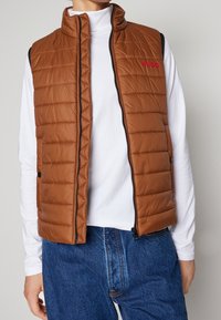 Braune gesteppte Weste mit hohem Kragen, Frontreißverschluss und rotem „HUGO“-Logo. Verfügt über schwarze Seiteneinsätze und wird über einem weißen Oberteil getragen.