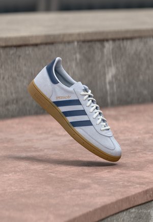 Λευκό αθλητικό παπούτσι Adidas Spezial με μπλε λωρίδες και σόλα καουτσούκ, αιωρούμενο πάνω από επιφάνεια από σκυρόδεμα.