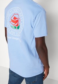 Tyrkysové bavlněné tričko s velkým grafickým potiskem červené a růžové růže se zelenými listy na zadní straně, potisknuté kruhovým textem kolem ní.