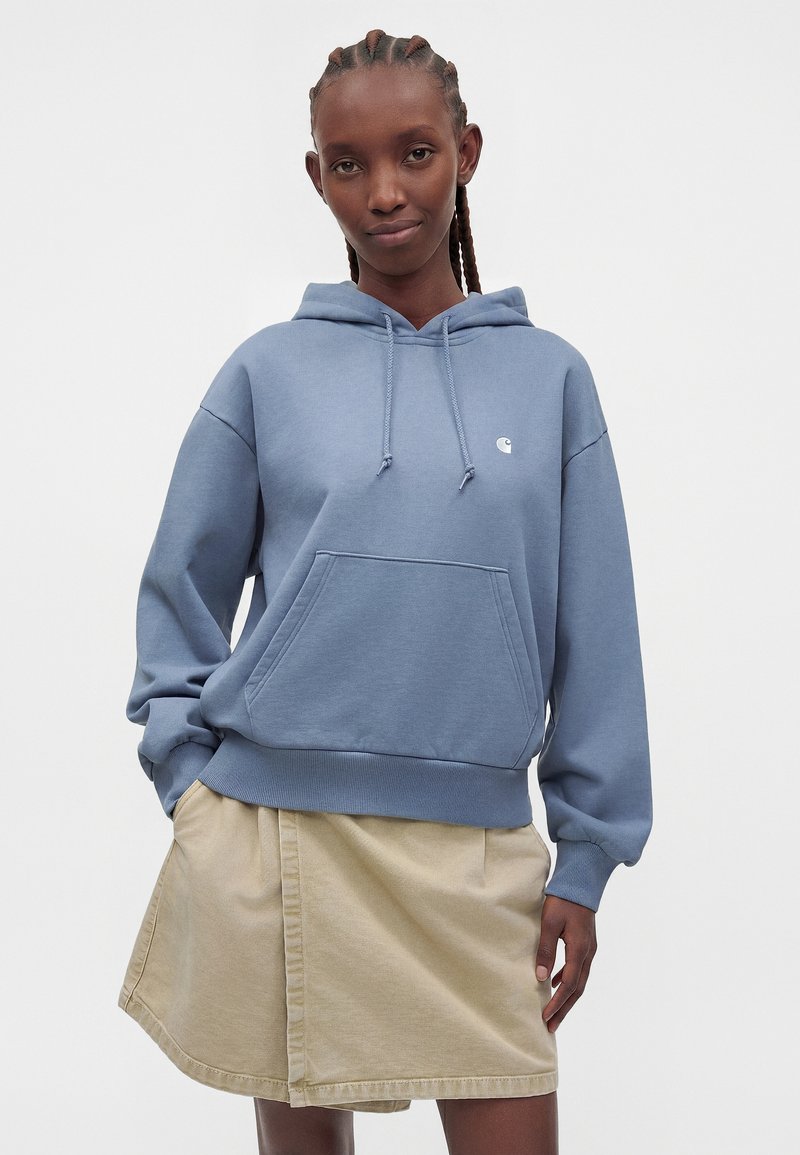 Young woman with braided hair wearing a light blue hoodie and beige skirt standing against a plain background.