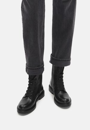 Black leather combat boots with a smooth finish, round toe, and lace-up front, paired with rolled gray denim pants. Textured fabric on the boots.