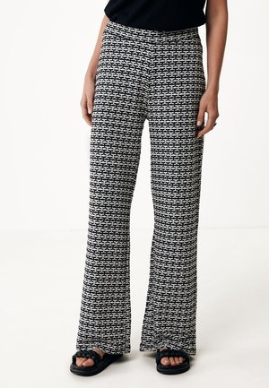 Femme portant un pantalon large taille haute à motif géométrique noir et blanc et des sandales noires, debout les mains détendues le long du corps.