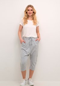 Kvinde med langt blond hår iført en hvid t-shirt, lysegrå capribukser med snøre og hvide sneakers, stående op ad en ensfarvet væg.