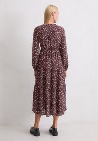 Długa, burgundowa sukienka w kwiaty z warstwowym fasonem, długimi rękawami i wcięciem w talii. Posiada małe, białe wzory kwiatowe i zaokrąglony dekolt.