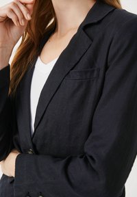 Tamsiai mėlynas lino švarkas su įpjovimu, vienu krūtiniu kišeniu ir mygtukų užsegimu. Turi švelnų tekstūrą ir pritaikytą siluetą virš balto marškinėlio.
