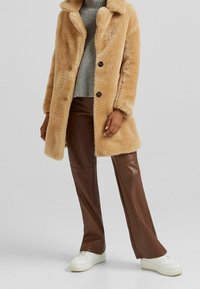 Abrigo de pelaje sintético beige con cuello de solapa, tres botones, usado sobre un suéter gris y pantalones de cuero sintético marrón, combinado con zapatillas blancas.