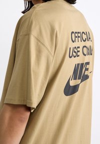 Camiseta de gran tamaño en color marrón, hecha de tejido suave, con estampado negro en la parte posterior que dice "USO OFICIAL SOLAMENTE" y "NIKE". Mangas cortas.