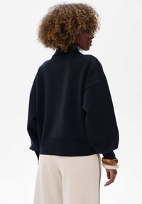 Czarny, oversize sweter z długimi, lekko bufkowymi rękawami. Dzianinowa faktura z prążkowanym wykończeniem. Dobrany z jasnobeżowymi spodniami i drewnianymi bransoletkami.