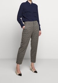 Žena ve tmavě modré blůze s dlouhým rukávem, vysokými kostkovanými kalhotami a béžovými lodičkami s černými špičkami, stojí s jednou rukou v kapse.