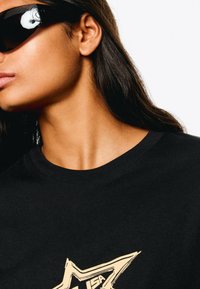 Black cotton t-shirt with a gold star graphic and text design. Model wears dark sunglasses, hair straight, with a smooth texture and neutral complexion.