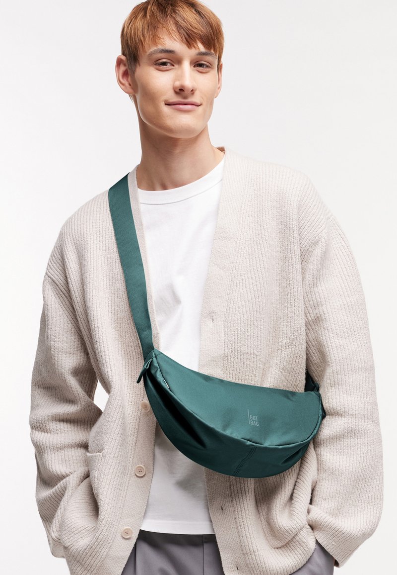 Junger Mann, der einen beigen Cardigan und ein weißes Hemd mit einer grünen Umhängetasche trägt, steht vor einem schlichten hellen Hintergrund.