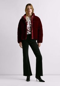 Rode velvet gewatteerde jas met een hoge kraag, gecombineerd met een wit grafisch T-shirt met lipafdrukken en zwarte uitlopende broek. Zwarte hakken.