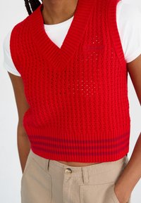 Rode gebreide mouwloze trui met een v-hals, voorzien van een textuurpatroon en een geribbelde zoom met een gestreept accent. Draagt over een wit shirt.