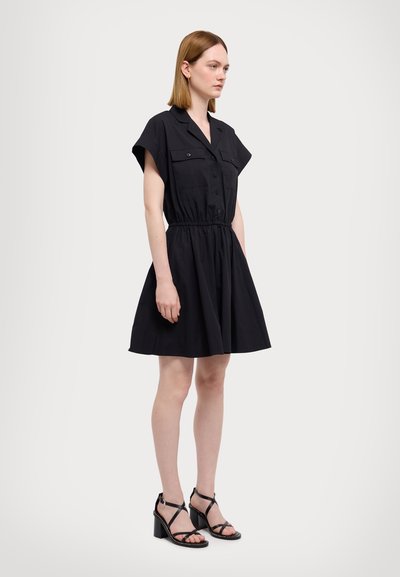 Robe noire à manches courtes avec deux poches poitrine, taille cintrée et jupe évasée. Associée à des sandales à talons noires à lanières.