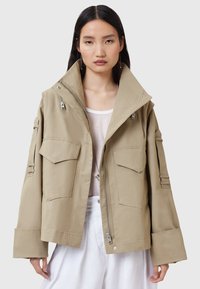 Giacca oversize khaki con doppie tasche sul petto, cerniere laterali e colletto alto. Realizzata in tessuto liscio, presenta maniche ampie e una vestibilità rilassata.