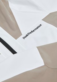 Weiße und beige Jacke mit geometrischem Design, schwarzem Reißverschluss und in schwarzem Stick auf dem Stoff aufgesticktem "PeakPerformance"-Logo.