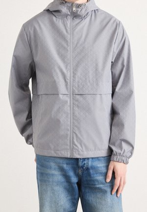 Personne portant une veste à capuche gris clair avec une fermeture éclair à l'avant et des poignets élastiques, associée à un jean en denim bleu.