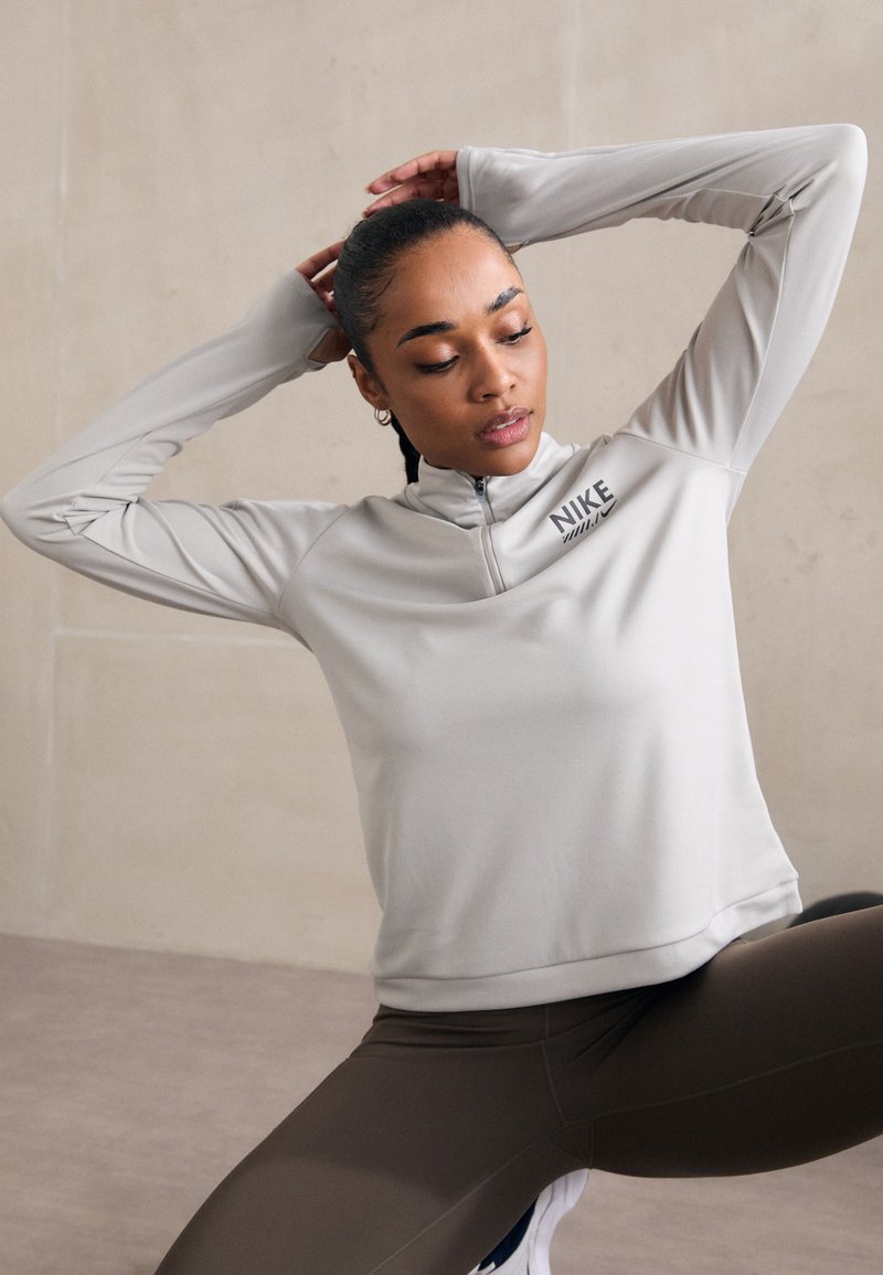 Lysegrå Nike-hoodie med halvlynlås, lange ærmer og en glat tekstur. Parret med mørkebrune leggings, der har et minimalt logo.