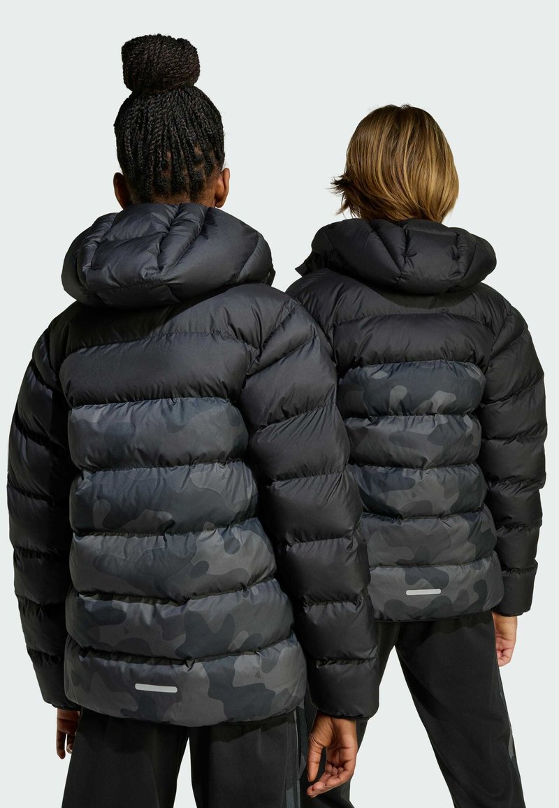 Les vestes matelassées noires avec capuchon présentent des sections en camouflage sombre contrastantes, un design matelassé et des accents réfléchissants le long de l'ourlet.