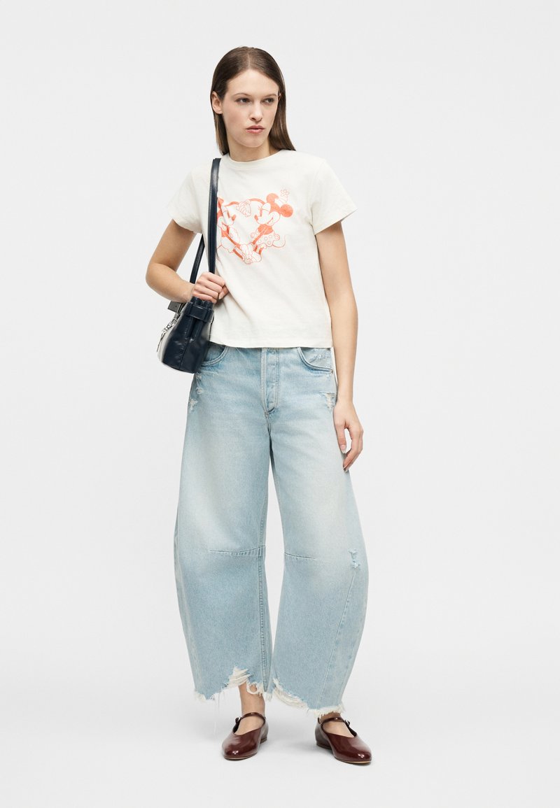 Frau trägt ein weißes T-Shirt mit Mickey-Mouse-Aufdruck, hellblaue, weit geschnittene Jeans mit Used-Effekten, braune Mary-Jane-Schuhe und eine schwarze Umhängetasche.