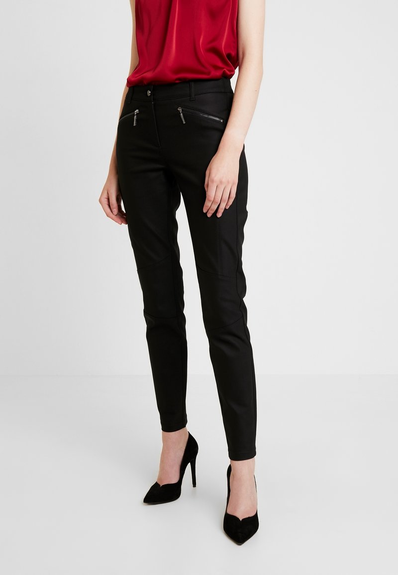 Femme portant un pantalon noir slim avec des poches zippées à l'avant, un haut rouge sans manches et des chaussures à talons noires, debout devant un fond uni.