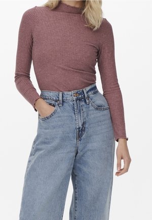 Vrouw draagt een mauve langärmelig geribd shirt, ingestopt in een hooggetailleerde, lichtblauwe wijde broek, met één hand in de zak.