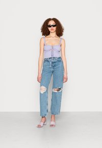 Top cropped em lavanda com detalhes de laço, jeans rasgados de cintura alta em azul claro e sandálias block heel em lavanda, contra um fundo simples.