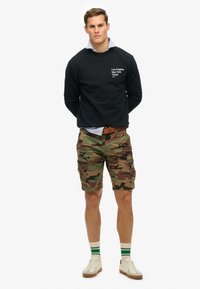 Sudadera negra con texto impreso, pantalones cortos cargo de camuflaje, calcetines a rayas blancas y verdes, y zapatillas beige. Estilo urbano y casual.