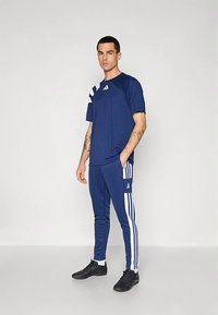 Marineblå sportsskjorte med hvite striper og logo, kombinert med matchende bukser med stripes på siden. Stoffet virker glatt og lett.