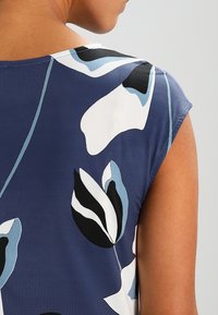 Haut bleu à fleurs avec manches courtes, présentant des motifs de feuilles noires et blanches. Tissu lisse avec une coupe ample et une couture d'épaule arrondie.