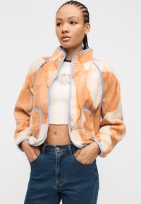 Νεαρή γυναίκα με πλεγμένα μαλλιά, ντυμένη με πορτοκαλί και μπεζ φλις ζακέτα με μπλε περιθώριο, λευκό κορμάκι και μπλε τζιν, με τα χέρια στις τσέπες.