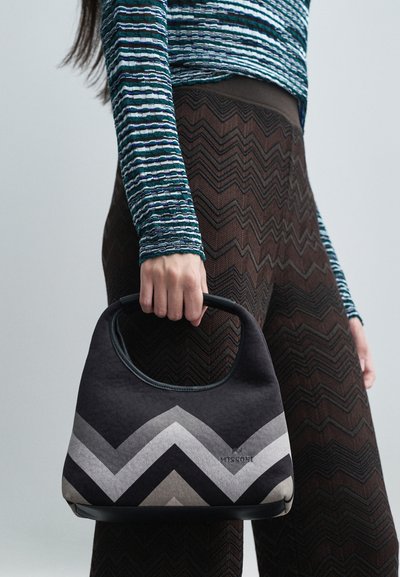 Άτομο που κρατάει μια τσάντα σε σχέδιο chevron μαύρου και γκρι χρώματος, φορώντας παντελόνι καφέ με σχέδιο ζιγκ-ζαγκ και μπλε ριγέ μπλούζα μακρύ μανίκι.