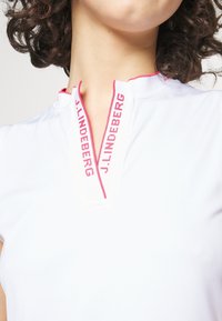 Vit polo med halvdragkedja, prydd med röd "J. Lindeberg"-logotyp på kragen. Slätt tyg och figurnära design.