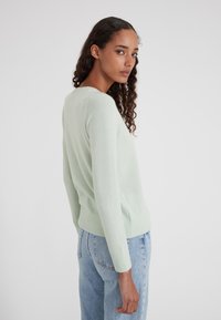 Donna con capelli ricci, che indossa un maglione verde chiaro e jeans blu, girata leggermente di lato su uno sfondo bianco semplice.