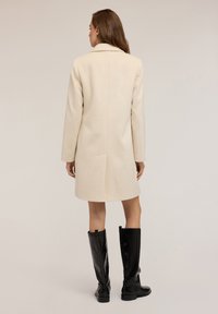 Donna che indossa un cappotto crema fino al ginocchio con uno spacco posteriore, abbinato a stivali neri al ginocchio decorati con borchie verticali.