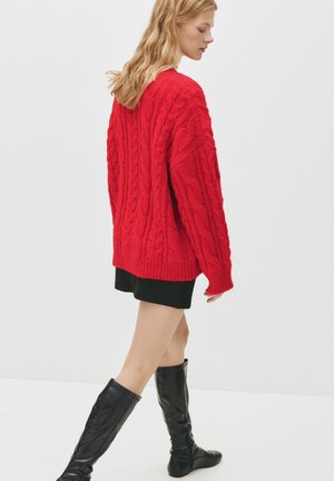 Czerwony sweter z dzianiny w warkocze o luźnym kroju, z ściśniętymi mankietami i dołem. W zestawie z czarną mini spódnicą i czarnymi kozakami do kolan.